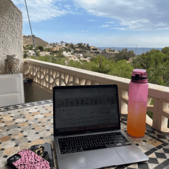 Photo of a laptop and a pink bottle on a table on a balcony in Spain. In the background, you can see trees, blue skies, and the sea.