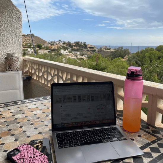 Foto van een laptop en roze fles op een tafeltje op een balkon in Spanje. Op de achtergrond zie je bomen, blauwe lucht en de zee