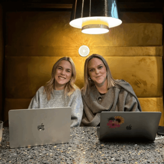 Two colleagues smiling at the camera. They are sitting at a table with their laptops, and a lamp is hanging above them.