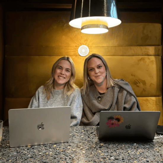 Twee collega's kijken lachend de camera in. Ze zitten aan een tafel met hun laptops en er hangt een lamp boven hun