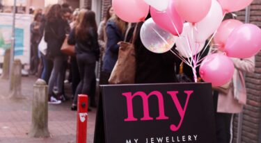 De opening van een My Jewellery Boutique winkel die feestelijk gevierd wordt. Je ziet op de voorgrond een uitklapbord met het logo en roze ballonnen en op de achtergrond zie je mensen in de rij staan aan de buitenkant van de winkel.