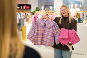 My Jewellery collega in de Boutique winkel die twee kledingstukken omhoog houdt voor een klant met stylingadvies en lacht naar de klant