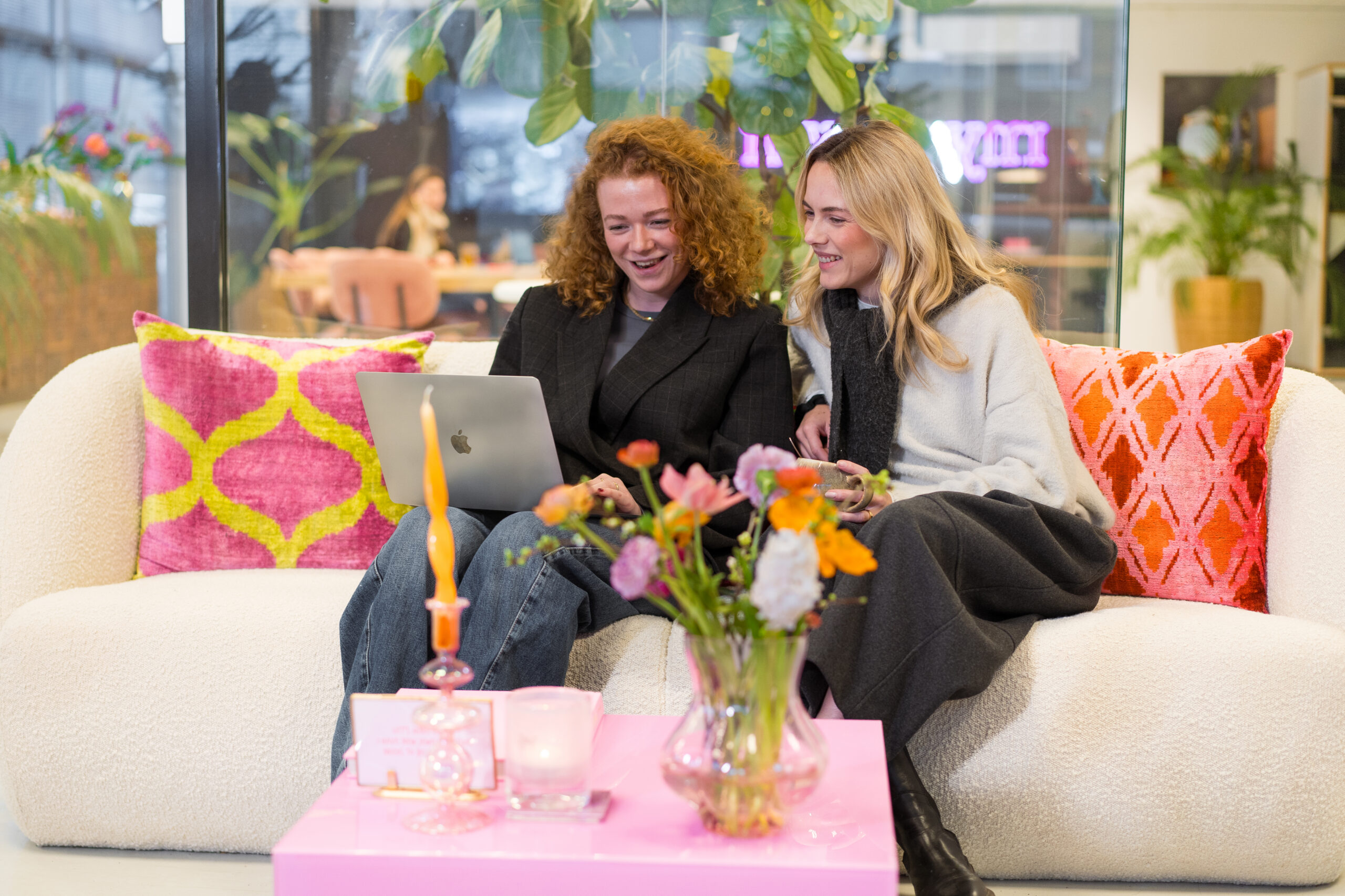 Zwei Recruiterinnen sitzen auf einem beigen Bouclé-Sofa mit rosa und roten Kissen und besprechen etwas gemeinsam. Sie schauen zusammen auf einen Laptop und lachen miteinander.
