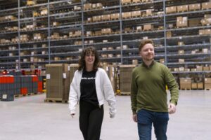 Twee My Jewellery collega's in My Warehouse in 's-Hertogenbosch die lopen door het magazijn en de stellages met dozen erop. Ze kijken trots vooruit en lachen.