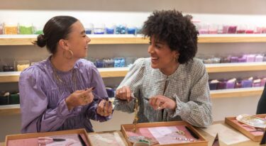 Twee collega's zijn bezig met het maken van sieraden in het My Jewellery Boutique Atelier winkel. Ze lachen naar elkaar terwijl ze dit doen. De ene collega houdt haar sieraad omhoog terwijl de andere collega met twee tangetjes bezig is aan haar ketting. Op de achtergrond zie je een aantal planken die vol staan met kleine bakjes met kralen in alle kleuren van de regenboog