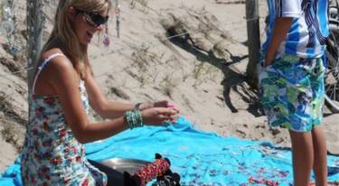 Sharon Hilgers, de founder en eigenaresse van My Jewellery, die op een blauw kleedje op het strand haar zelfgemaakte sieraden verkoopt aan een jongetje in een voetbalshirt en zwembroek. Ze houdt een sieraad uit naar het jongetje en lacht naar hem. Je ziet het blauwe kleed vol liggen met sieraden en daar achter het zand.