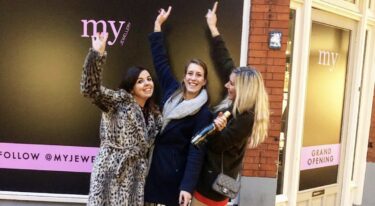 Sharon Hilgers, de founder en eigenaresse van My Jewellery, die de allereerste Boutique winkel in 's-Hertogenbosch viert samen met twee collega's (Jasmijn en Megan) met een fles champagne. Ze staan alledrie met 1 arm in de lucht en hebben grote glimlachen op hun gezicht. Ze staan voor de etalage van de nieuwe winkel