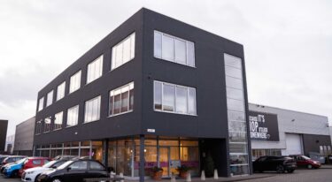 De buitenkant van het My Jewellery Hoofdkantoor in 's-Hertogenbosch. Het is een donkergrijs pand met drie verdiepingen en verschillende ramen. Aan de rechterkant zie je een magazijn en aan de linkerkant zie je geparkeerde auto's staan