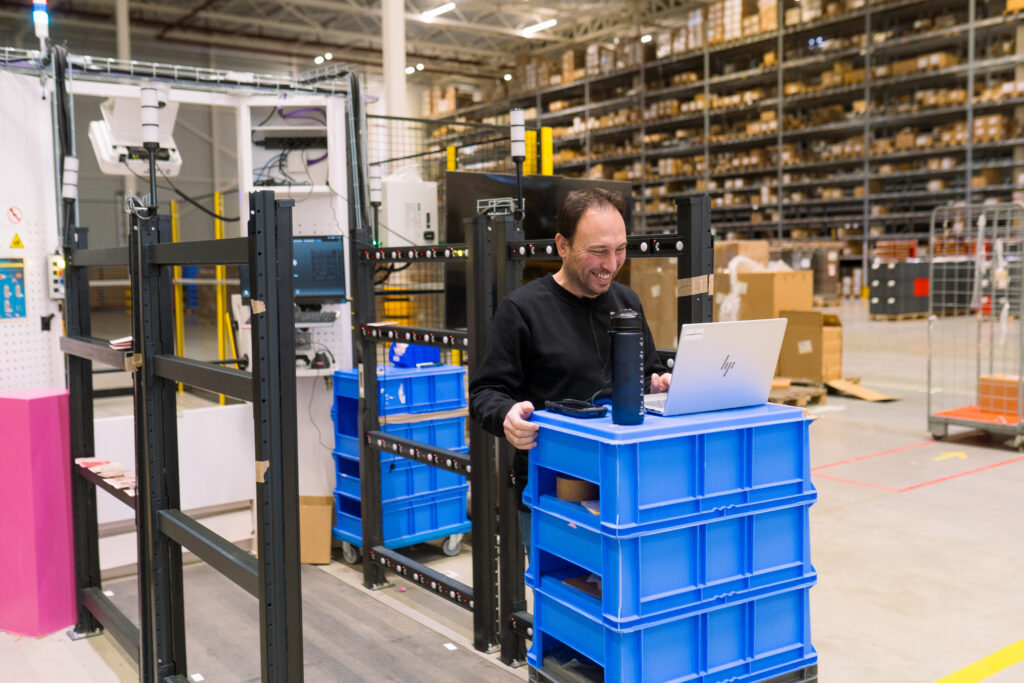 Een senior voorraad beheerder die lachend op zijn laptop kijkt en omringd is door dozen in het logistieke magazijn van My Warehouse en de Geek robots