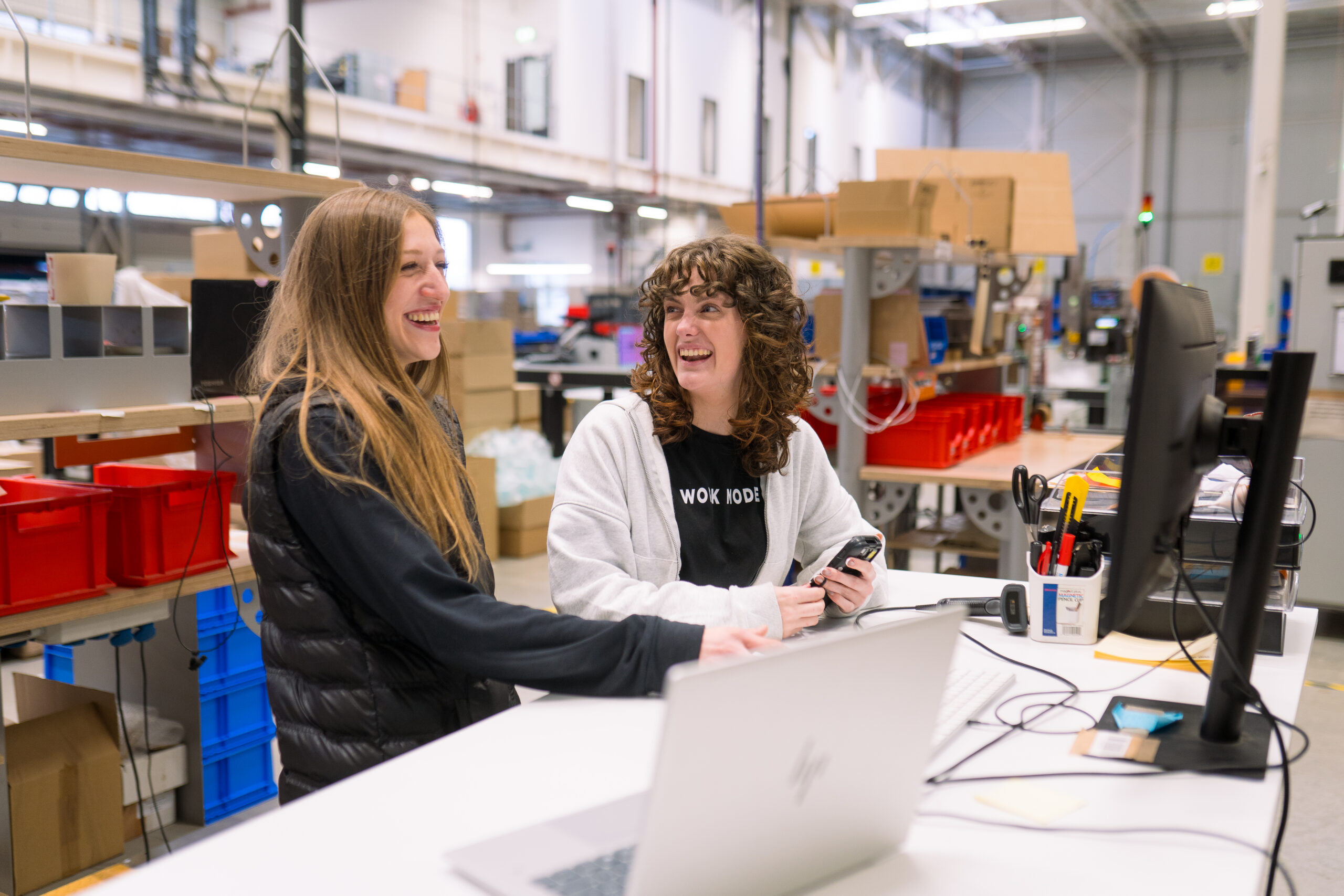 Twee logistieke medewerkers van My Warehouse, het magazijn van My Jewellery, die samen lachen achter een bureau met computers, met een scanner in de hand en omringd door dozen