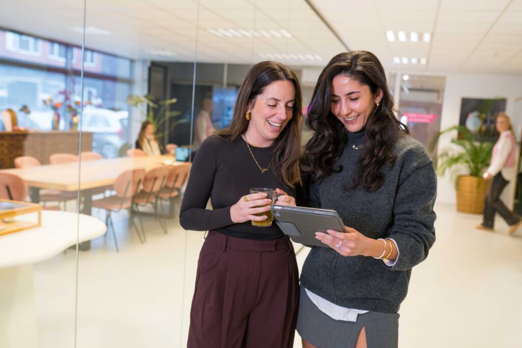 Twee collega's op het My Jewellery Hoofdkantoor in 's-Hertogenbosch staan langs een meeting ruimte en kijken samen op een tablet terwijl ze glimlachen. Op de achtergrond zie je een collega aan het werken en een andere collega aan het rondlopen. In de meetingruimte staan roze stoelen en bloemen en planten.
