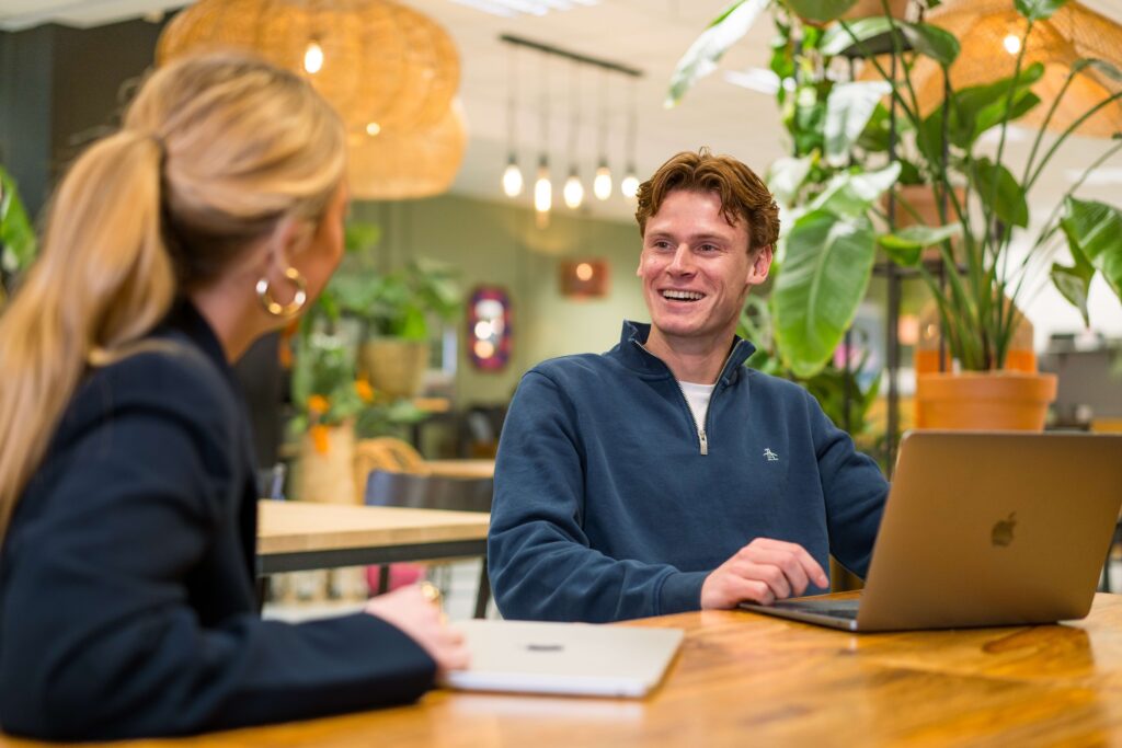 Een lachende collega in een blauwe polo van het My Jewellery Hoofdkantoor in 's-Hertogenbosch die achter zijn laptop zit en lachend naar een collega kijkt die je van de achterkant ziet. Op de achtergrond zie je diverse planten en rieten lampen hangen