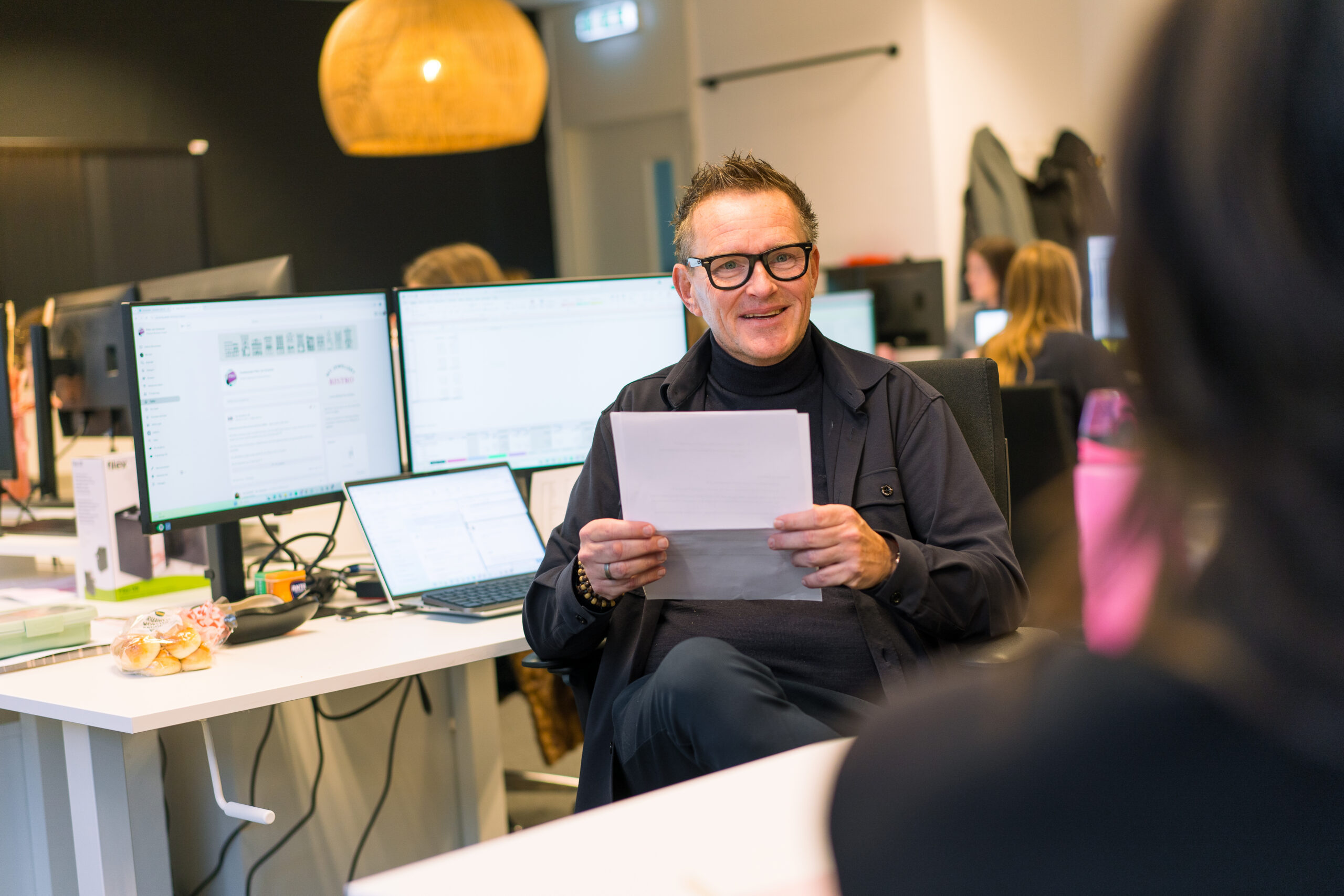 Business Controller van het My Jewellery Hoofdkantoor in 's-Hertogenbosch aan het praten met een andere collega met een glimlach op zijn gezicht. In zijn handen heeft hij papieren vast en op de achtergrond zie je zijn beeldschermen en collega's aan het werk