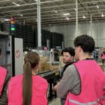 Studenten van de Avans Hogeschool op bezoek bij My Warehouse van My Jewellery in 's-Hertogenbosch. Er staan een aantal studenten met roze veiligheidshesjes te luisteren naar een collega bij de verpakkingsmachines.