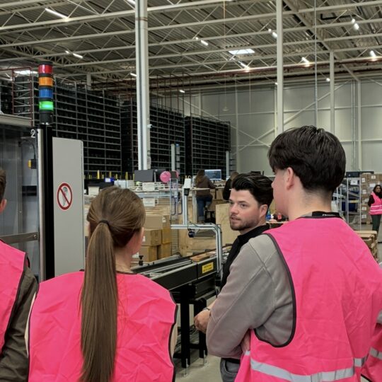 Studenten van de Avans Hogeschool op bezoek bij My Warehouse van My Jewellery in 's-Hertogenbosch. Er staan een aantal studenten met roze veiligheidshesjes te luisteren naar een collega bij de verpakkingsmachines.