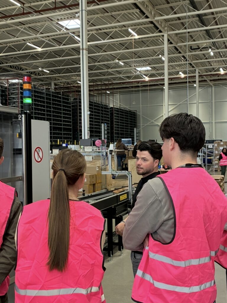 Studierende der Avans Hogeschool besuchen das My Warehouse von My Jewellery in ’s-Hertogenbosch. Mehrere Studierende mit rosa Sicherheitswesten hören einer Kollegin oder einem Kollegen bei den Verpackungsmaschinen aufmerksam zu.