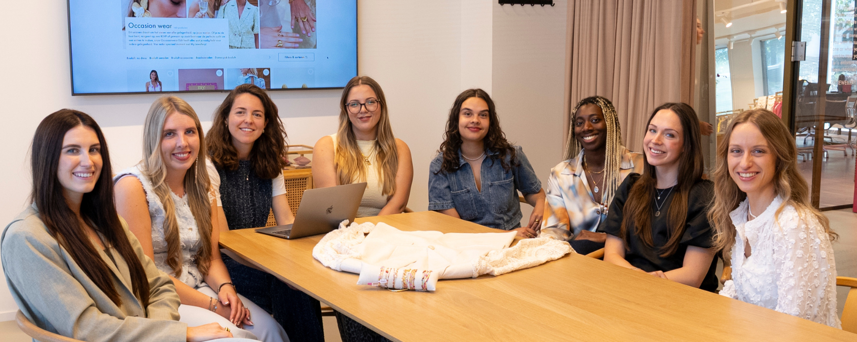 Teamfoto van het content team. Er zitten 8 vrouwen aan een tafel in een vergaderruimte die de camera inkijken en lachen. Op de achtergrond zie je een tv-scherm met de webshop en op de tafel ligt een kledingstuk.