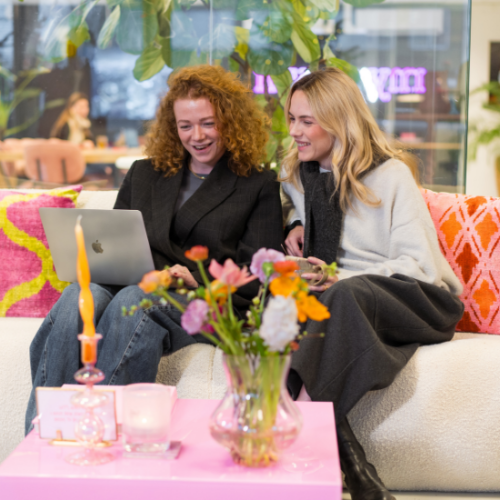 Zwei Recruiterinnen sitzen auf einem beigen Bouclé-Sofa mit rosa und roten Kissen und besprechen etwas gemeinsam. Sie schauen zusammen auf einen Laptop und lachen miteinander.