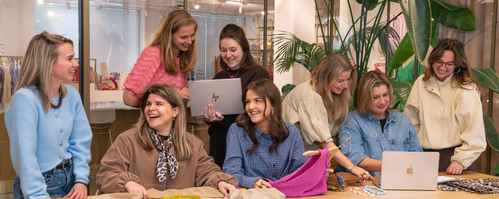 Groepsfoto van het Inkoop (productie) team op het Hoofdkantoor van My Jewellery in 's-Hertogenbosch. Er zijn acht mensen in een meetingruimte aan het overleggen met elkaar in kleinere groepjes. Op de tafel staan laptops en liggen en aantal producten.