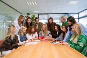 Gruppenfoto des HR-Teams in der My Jewellery Zentrale in ’s-Hertogenbosch. Es sind 13 Kolleginnen und Kollegen in einem Besprechungsraum, die um einen Tisch sitzen und stehen. Sie schauen gemeinsam auf einige Broschüren auf dem Tisch und lachen miteinander.