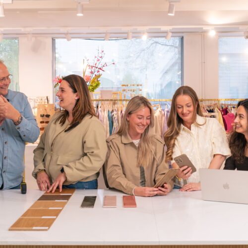 Groepsfoto van het Retail Backoffice team op het Hoofdkantoor van My Jewellery in 's-Hertogenbosch. Er staan vijf mensen achter een witte balie in de showroom. De linker twee kijken samen naar staaltjes van houten vloeren en tegels. Drie collega's daar rechts van overleggen over een tegel met een laptop erbij.