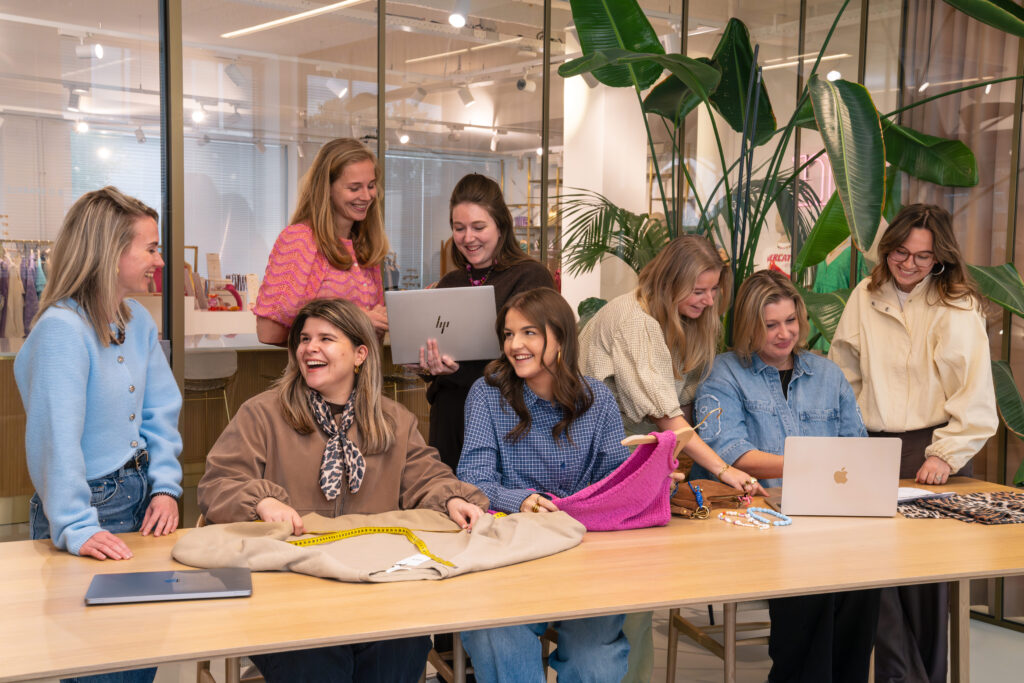 Groepsfoto van het Inkoop (productie) team op het Hoofdkantoor van My Jewellery in 's-Hertogenbosch. Er zijn acht mensen in een meetingruimte aan het overleggen met elkaar in kleinere groepjes. Op de tafel staan laptops en liggen en aantal producten.