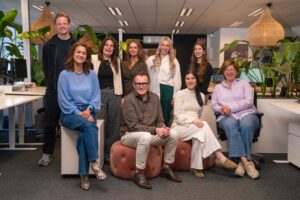 Groepsfoto van het Finance team op het Hoofdkantoor van My Jewellery in 's-Hertogenbosch. Er zijn negen mensen in een kantoortuin, waarvan sommige staan en sommige zitten op een bureaustoel of poef. Ze kijken lachend naar de camera.