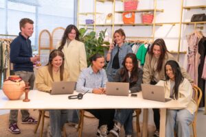 Groepsfoto van het Customer Care team op het Hoofdkantoor van My Jewellery in 's-Hertogenbosch. Er zijn acht mensen bij een tafel in de showroom. Drie mensen zitten aan de tafel te overleggen en eentje zit te bellen met een headset. Daarachter zie je drie collega's ook overleggen en de andere aan het bellen met haar telefoon.