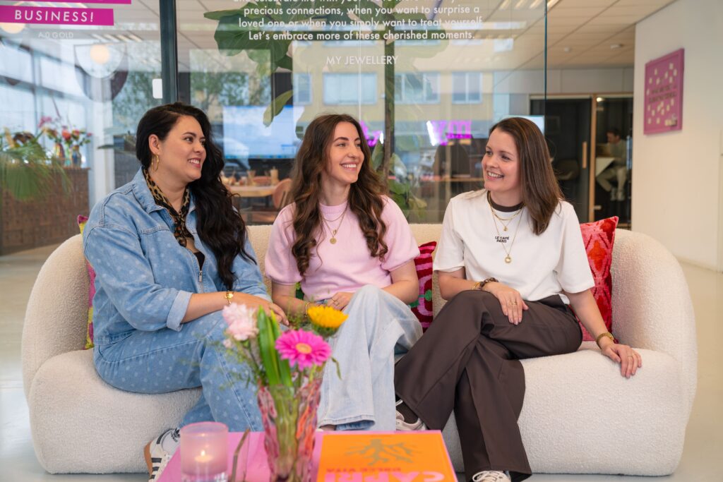 Groepsfoto van het Digital Merchandising team op het Hoofdkantoor van My Jewellery in 's-Hertogenbosch. Drie vrouwen zitten op een witte bank en lachen naar elkaar.