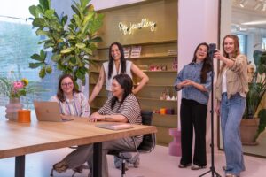 Gruppenfoto des Social-&-SEA-Teams in der My Jewellery Zentrale in ’s-Hertogenbosch. Drei Personen besprechen sich an einem Tisch in einem Besprechungsraum mit ihren Laptops. Rechts neben dem Tisch nehmen zwei Kolleginnen und Kollegen Content mit ihrem Smartphone und einem Stativ auf.