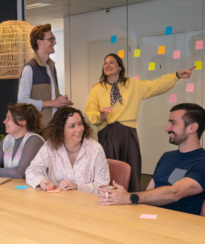 Groepsfoto van het Online Customer Experience team op het Hoofdkantoor van My Jewellery in 's-Hertogenbosch. Vier mensen zitten aan een tafel in een meetingruimte te overleggen. Daarachter staan drie collega's die naar post-its kijken die hangen op de glazen wand.