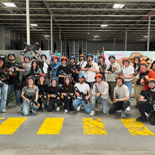 Teamphoto in My Warehouse in 's-Hertogenbosch. A group of employees with helmets on and lasergameguns in their hands laughing at the camera