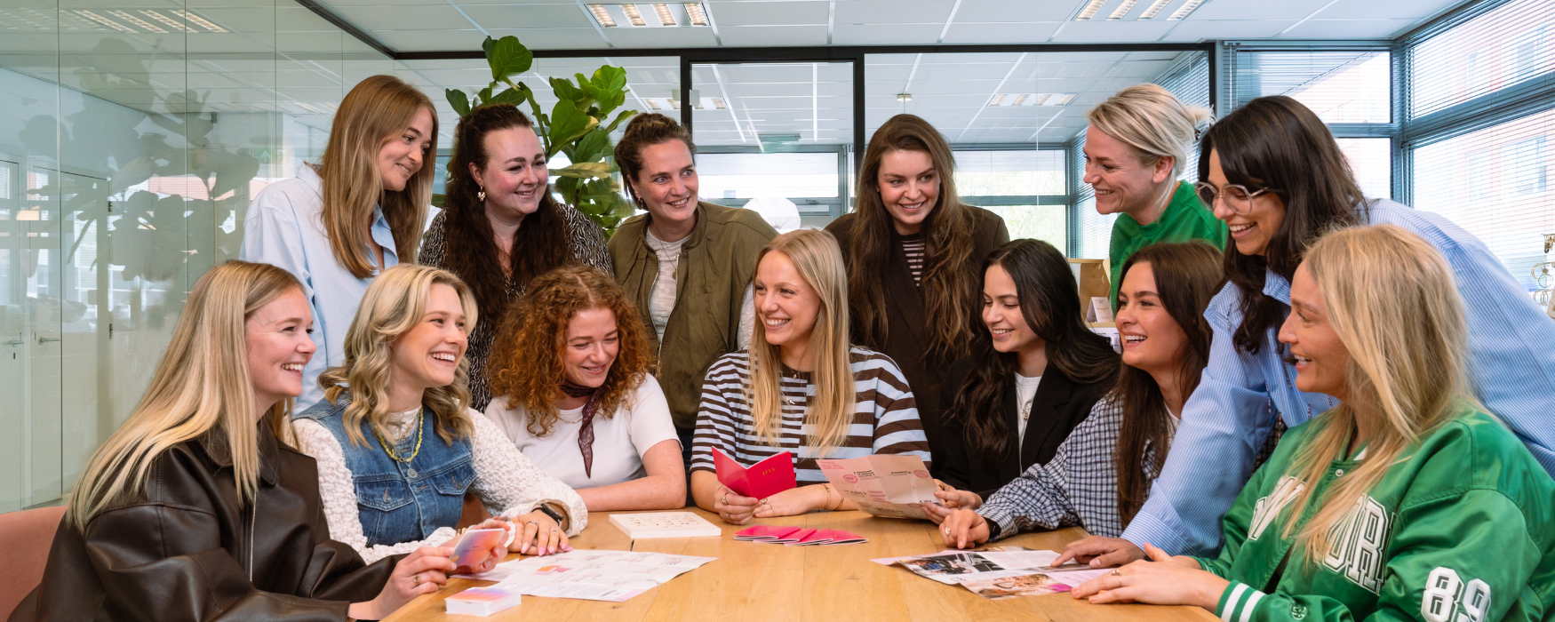 Groepsfoto van het HR team op het Hoofdkantoor van My Jewellery in 's-Hertogenbosch. Het zijn 13 collega's in een meetingruimte die om een tafel zitten en staan. Ze kijken naar een aantal boekjes op tafel en lachen naar elkaar.