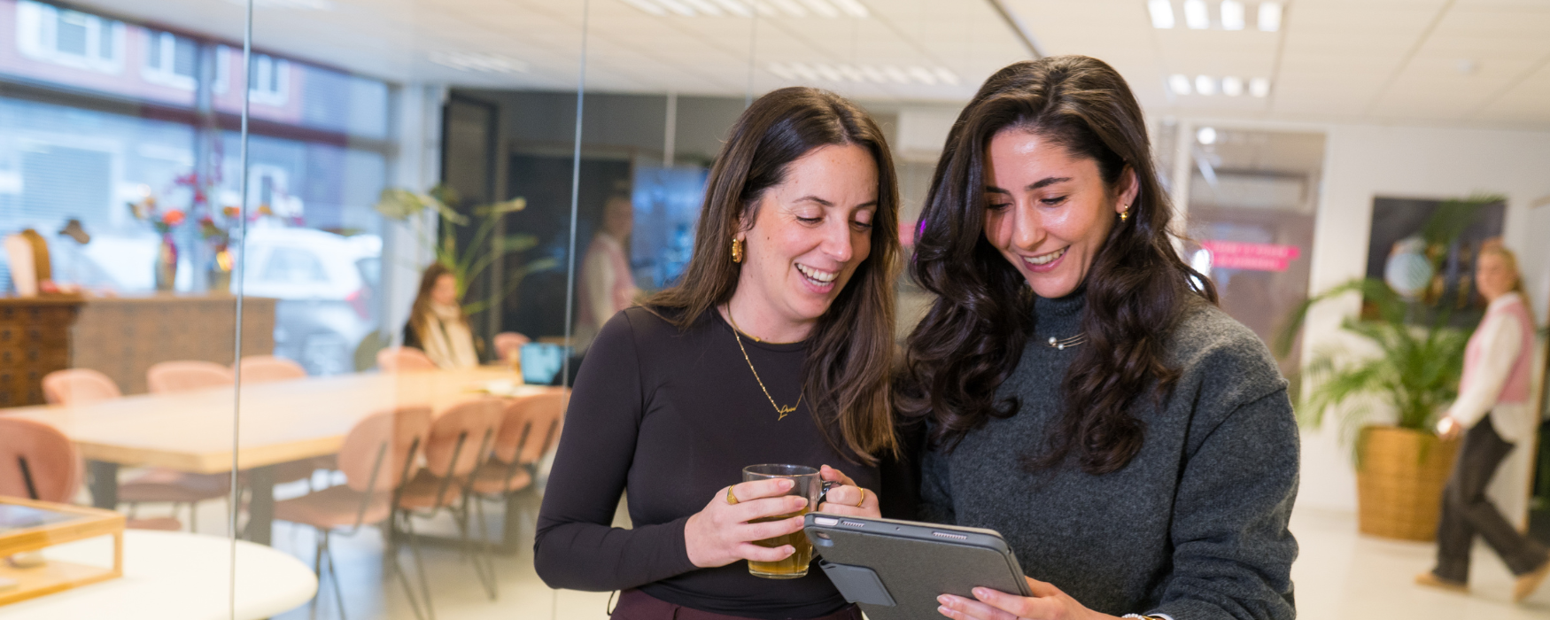 Twee collega's op het My Jewellery Hoofdkantoor in 's-Hertogenbosch staan langs een meeting ruimte en kijken samen op een tablet terwijl ze glimlachen. Op de achtergrond zie je een collega aan het werken en een andere collega aan het rondlopen. In de meetingruimte staan roze stoelen en bloemen en planten.
