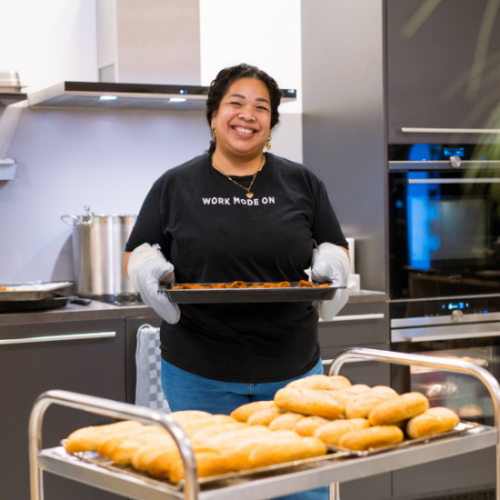 Catering beheerder van My Jewellery Hoofdkantoor in 's-Hertogenbosch in de Bistro kantine bezig met het voorbereiden van eten met een glimlach op haar gezicht. Ze heeft ovenwanten aan en draagt een ovenschaal met eten, voor haar staan schalen met broodjes en je ziet de soeppan op de achtergrond.