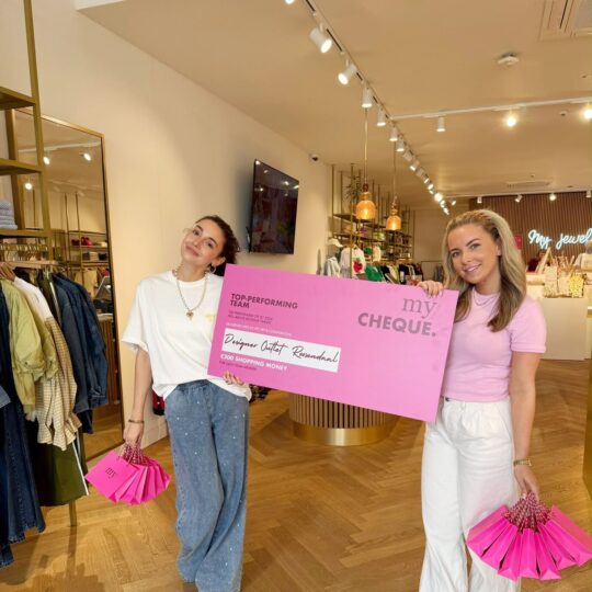 Deux femmes de la boutique My Jewellery du Designer Outlet Roosendaal sourient à la caméra, un chèque rose à la main