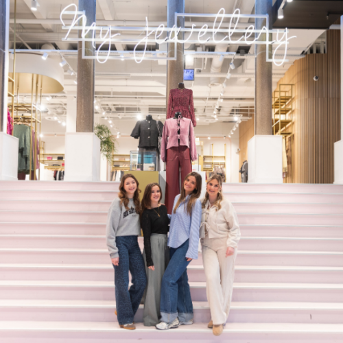 Vier collega's van de My Jewellery Boutique winkel in Eindhoven staan op een roze trap terwijl ze naar de camera lachen. In het midden van het beeld staan een drietal paspoppen op de trap en boven de trap hangt een groot logo van My Jewellery.